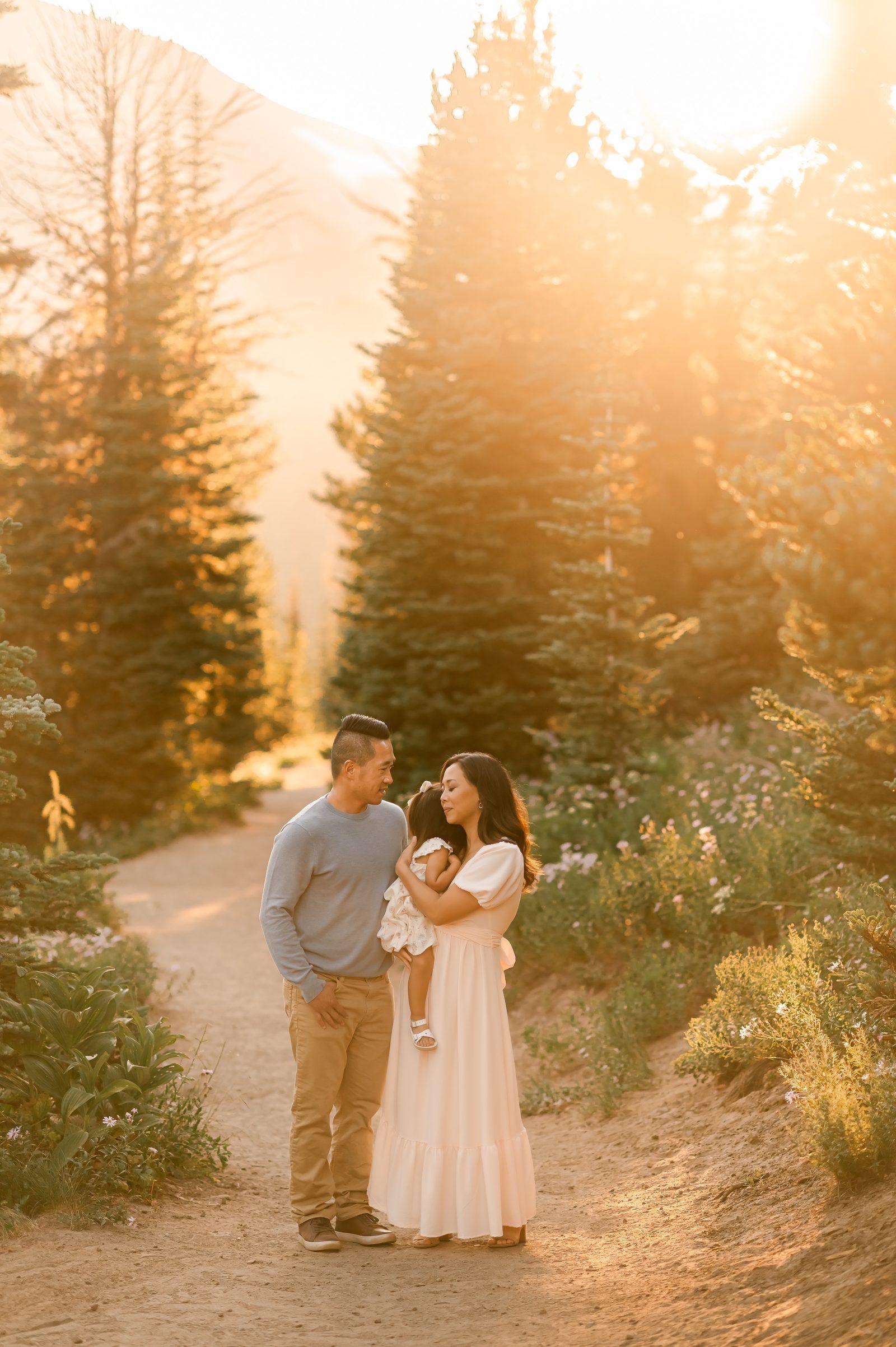 A couple on a mountain path at golden hour, holding a small child, with wildflowers and pines on either side