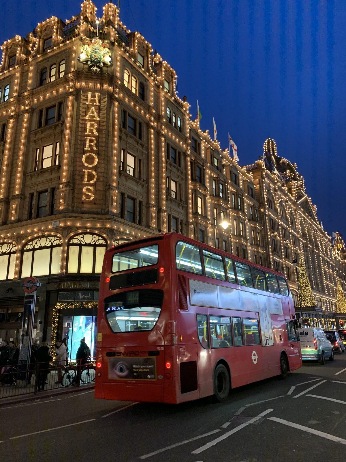 Harrods lit up at dusk with a red double-decker bus passing